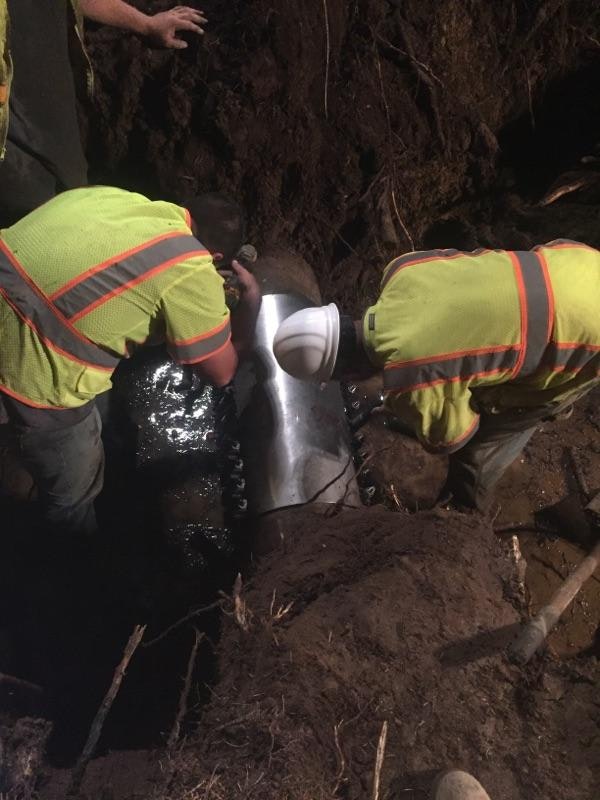 Two workers in high-visibility gear installing pipe fittings in a trench.