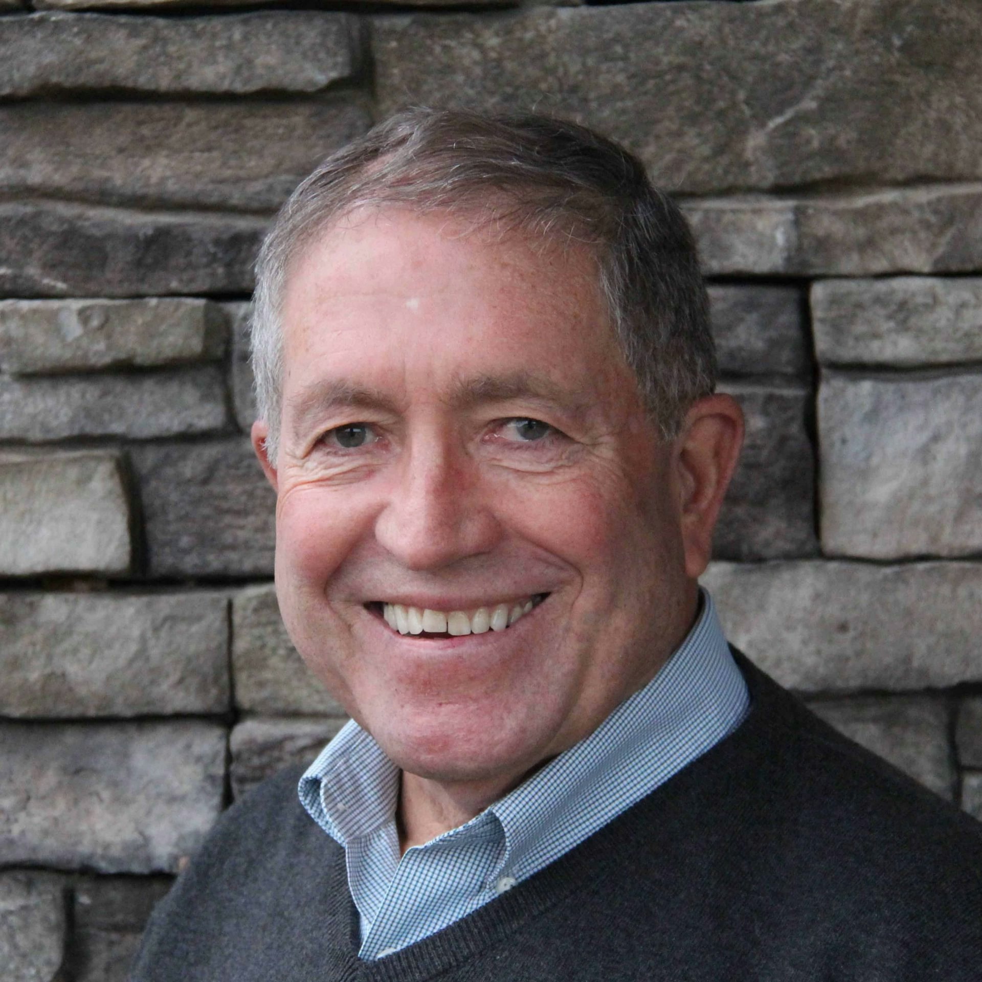 Smiling man with grey hair, wearing a sweater over a collared shirt, in front of a stone wall.