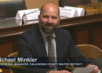 A smiling man at a desk with a nameplate and title in front of a microphone at a Calaveras County Water District meeting.