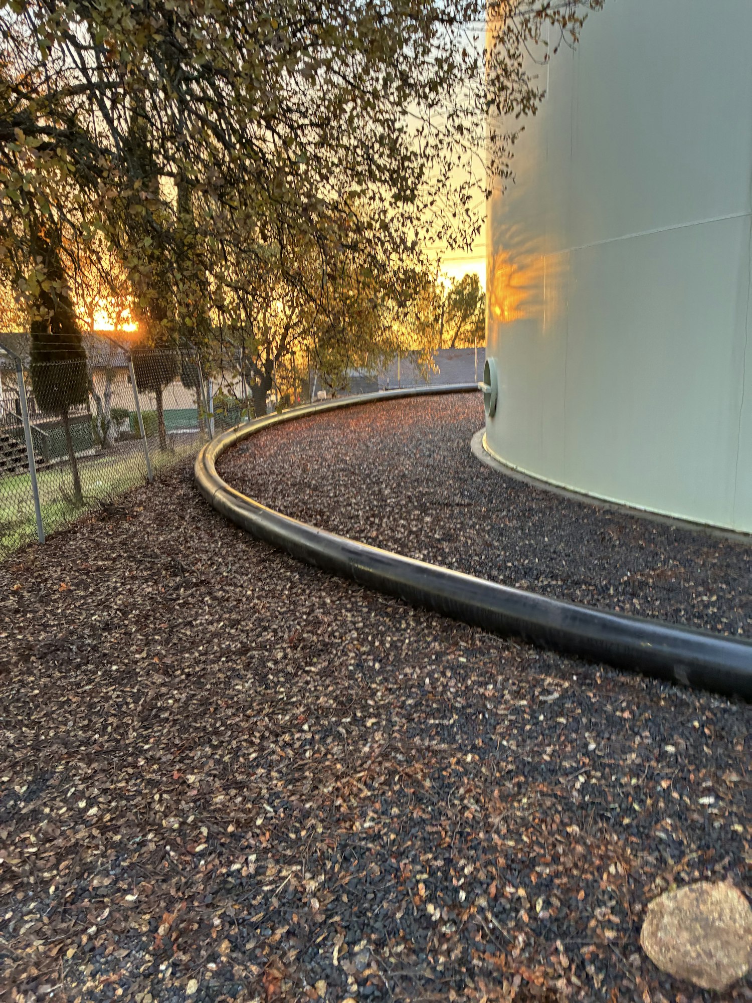 The image shows a curved black pipe, gravel ground, and a sunset background with trees and a fence.