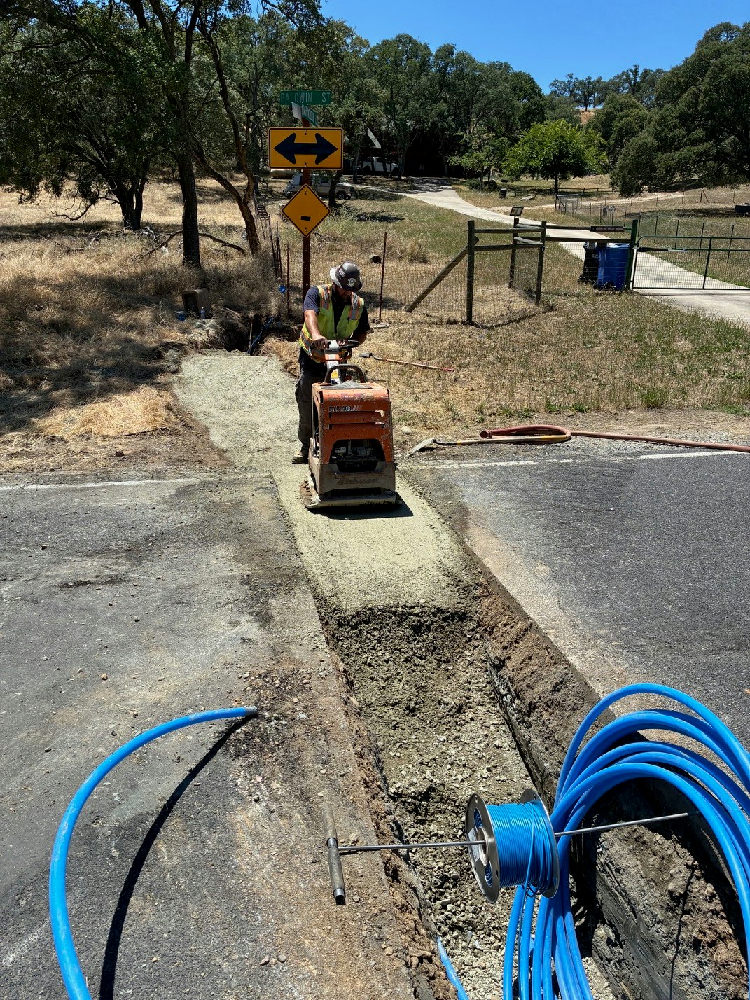 A worker operates machinery on a road, surrounded by tools and utility equipment, with trees and a road sign in the background.