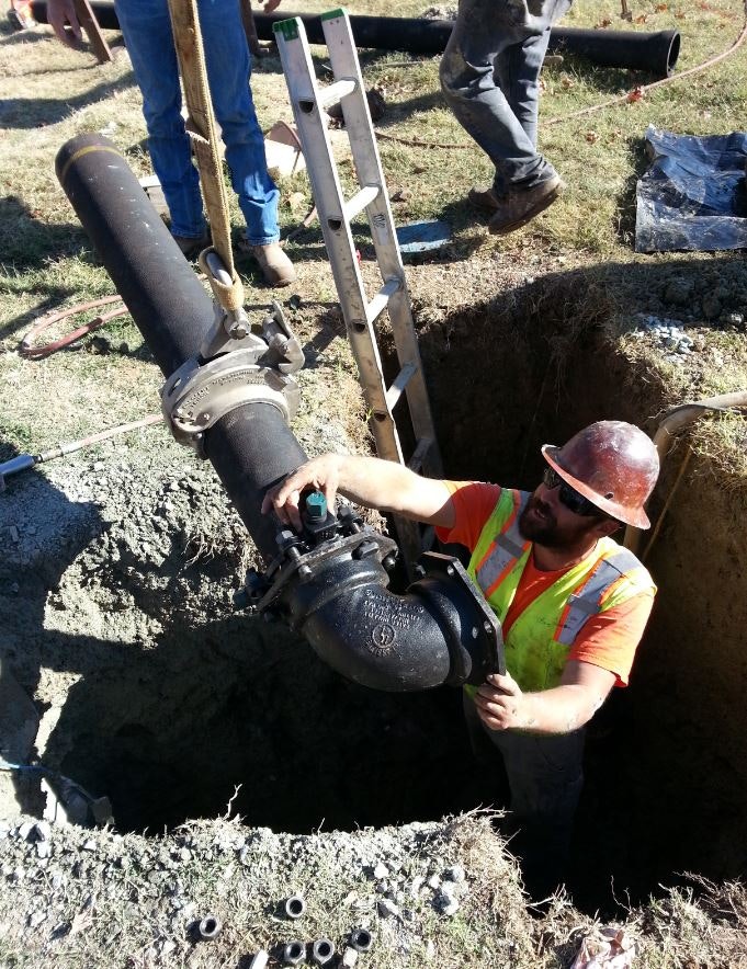 Worker in a trench installing a large pipe with tools and components around; others observe.