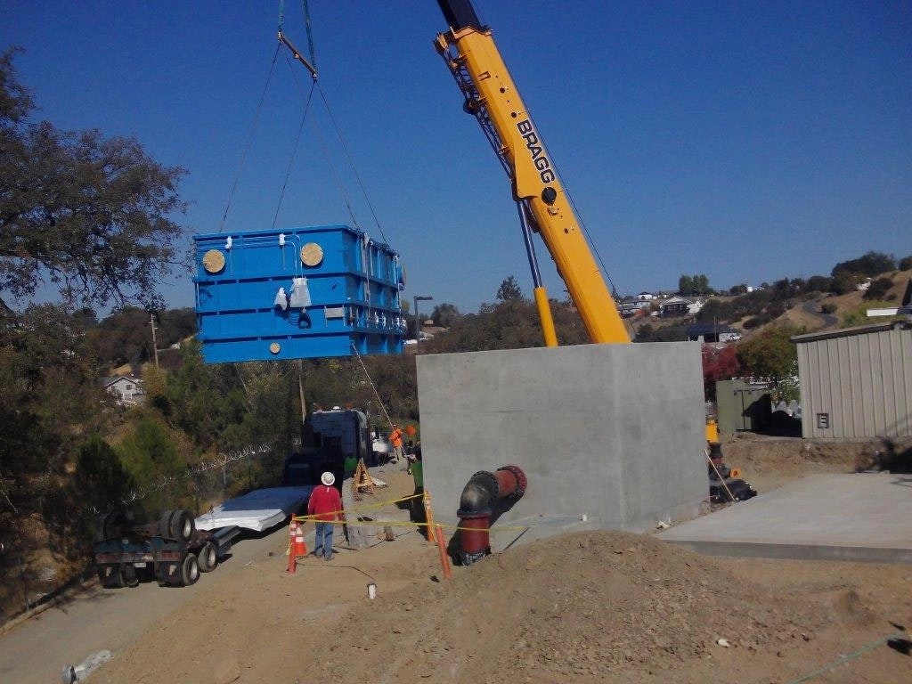 Construction site with a crane lifting a blue container, workers, concrete walls, clear sky, and distant houses.
