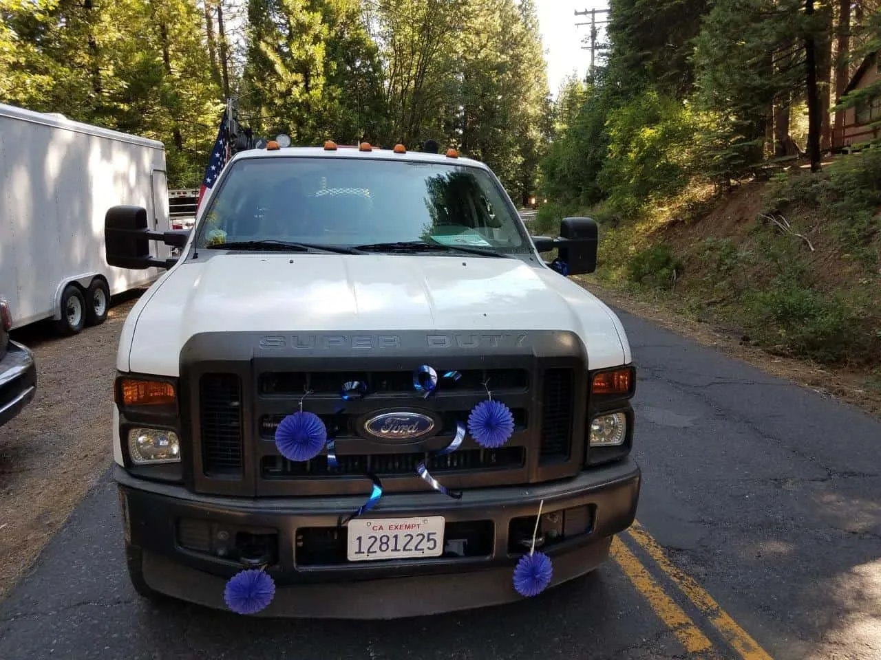 A Ford Super Duty truck with blue decorations on the grill, parked on a forest-lined road, with an American flag and trailer in the background.