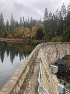A dam with a calm water reservoir surrounded by forested area. Cloudy sky above.