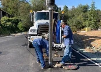 Two workers operating a drilling rig on a sunny road.