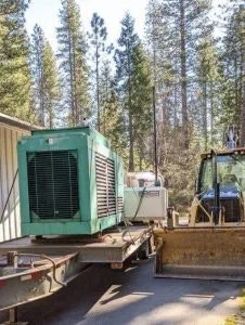 A generator on a trailer, a smaller unit beside it, and a backhoe, all parked in a wooded area.