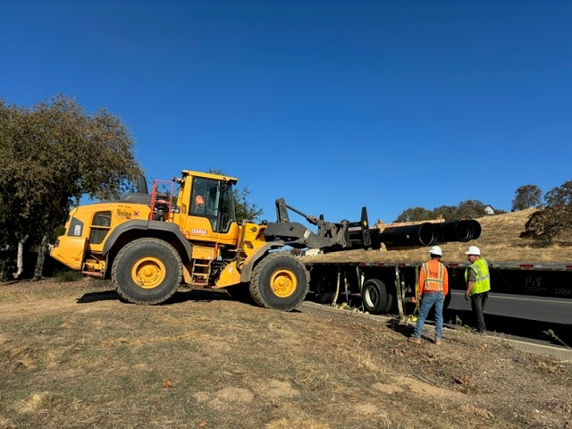 A yellow construction vehicle works near a road while two workers observe, with blue skies and trees in the background.