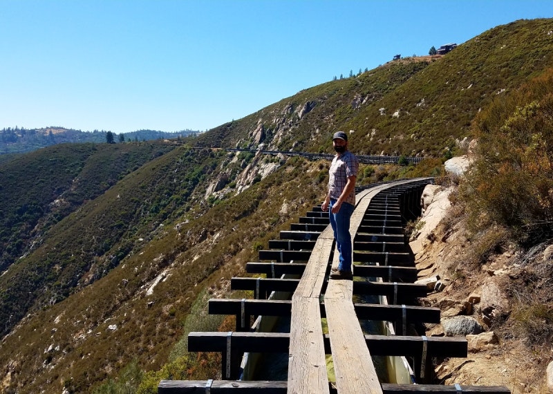 A person stands on a disused wooden railroad bridge in a mountainous area.