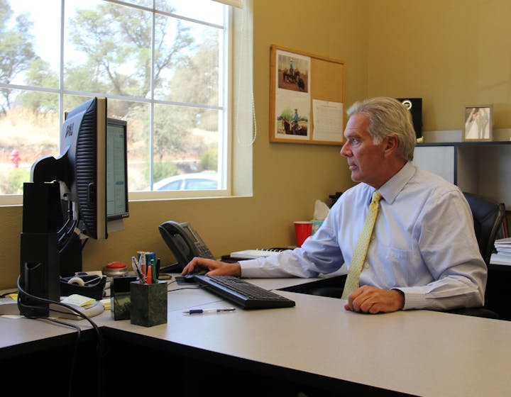 A man in a tie working at a desk with a computer, phone, and office supplies.