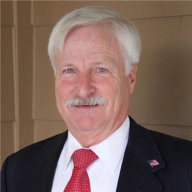 A smiling man with a white mustache, wearing a suit, red tie, and an American flag pin.