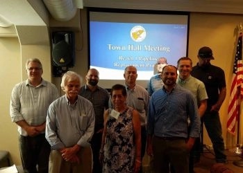 A group of people smiling in front of a screen displaying "Town Hall Meeting" with an American flag to the side.