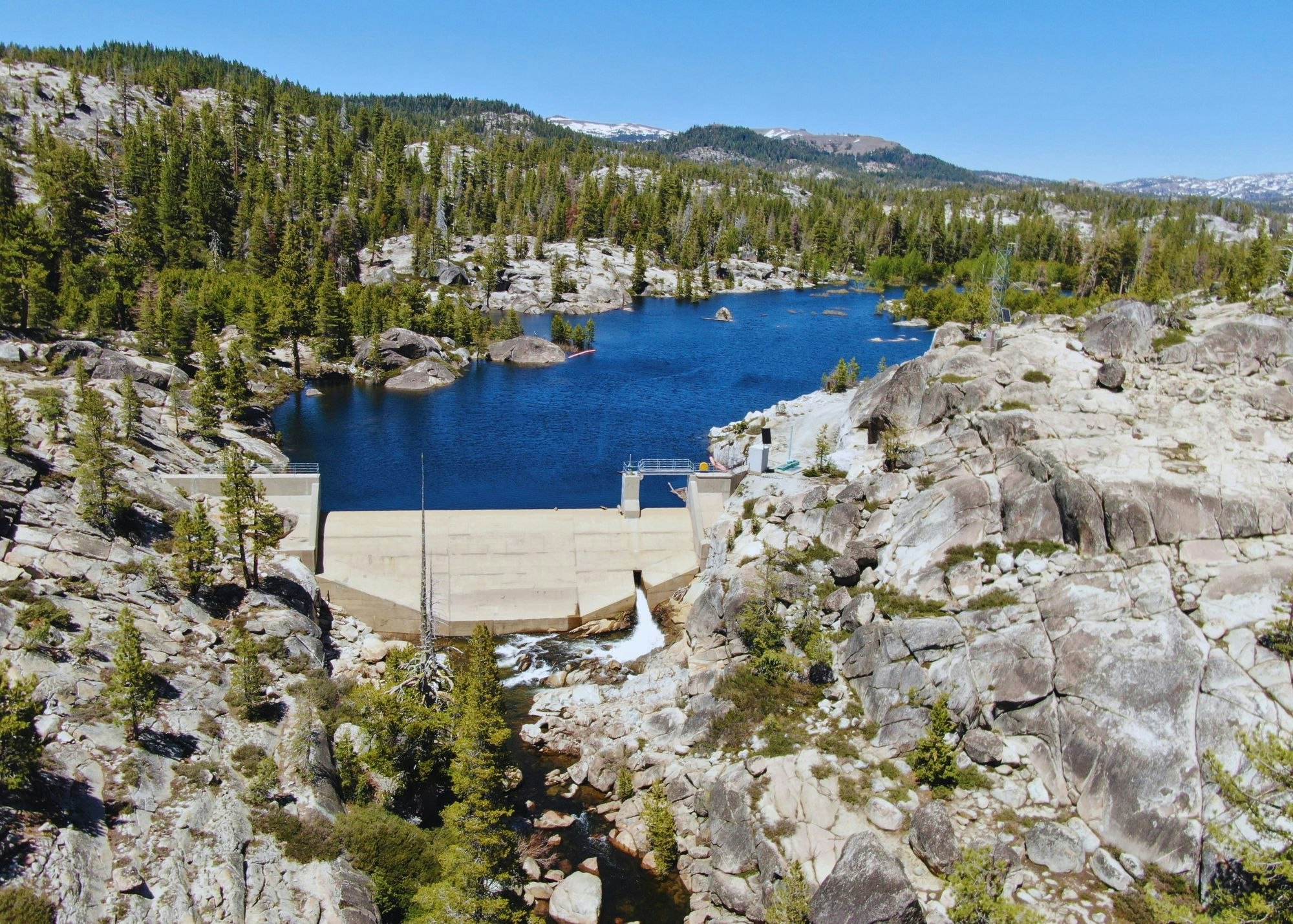 A dam with water flowing through mountains and a forest, under a clear blue sky.