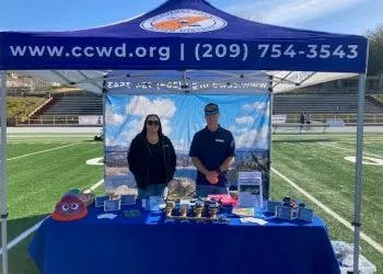 Two people at an informational booth with a banner, brochures, and various display items on a table.