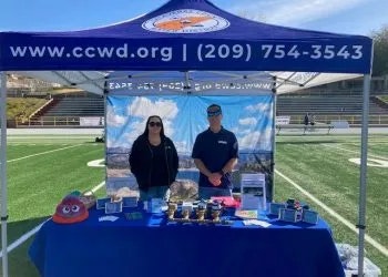 Two people at an informational booth with a banner, brochures, and various display items on a table.