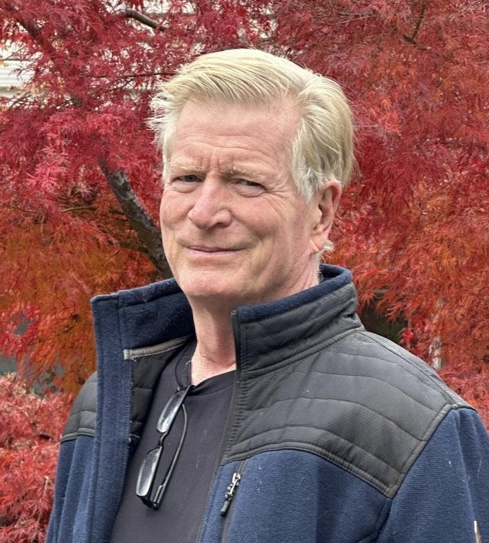 A person in a blue jacket stands in front of vibrant red foliage, smiling.