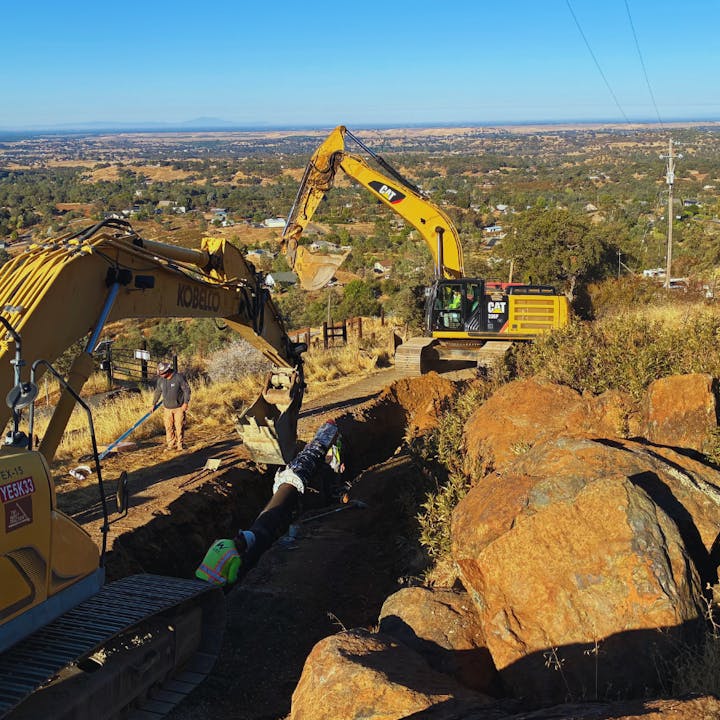 Construction site with two excavators and workers digging a trench in a rural area.