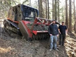 Two individuals posing next to a forest mulching machine in a wooded area.