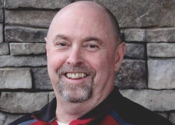 A smiling man with a mustache and goatee against a stone wall background.