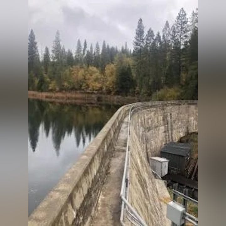 A dam wall beside a tranquil reservoir, surrounded by forested hills under an overcast sky.
