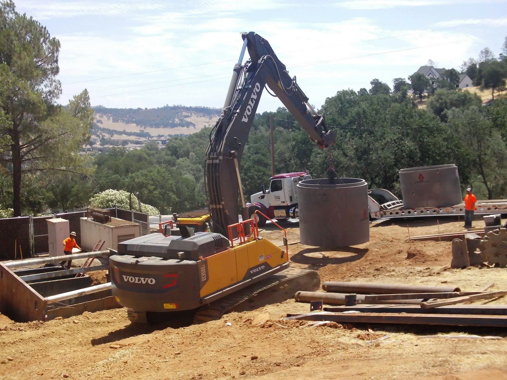 A Volvo excavator at a construction site with workers, material on the ground, and scenic hills in the background.