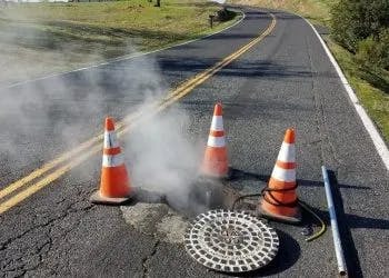 Steam rising from a manhole on a road with traffic cones and barriers around it.
