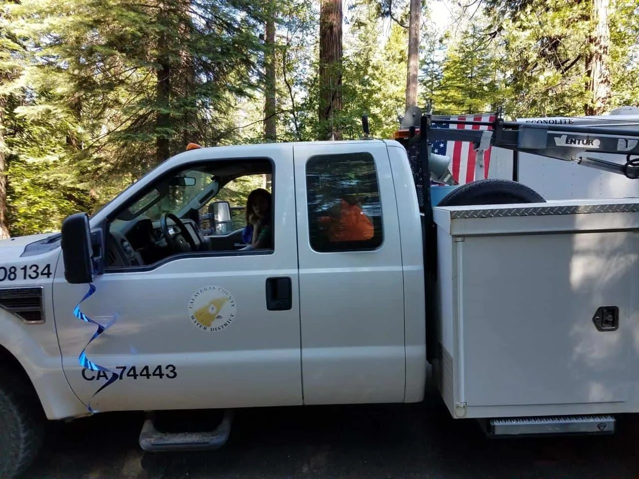 A white utility truck with California license plate and logo parked among trees, a person inside, and American flag decals.