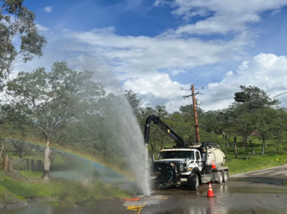 A truck spraying water with a rainbow visible in the mist, under a blue sky with clouds.