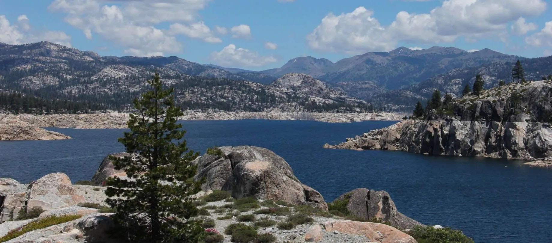 Mountainous landscape with a lake, rocky shores, pine trees, and fluffy clouds under a blue sky.