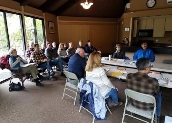A group of people seated around tables in a meeting or class, focusing attention on a person speaking.