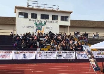 A group of people seated in stadium bleachers at "Home of the Bullfrogs, Dorroh Field," with event sponsor banners in front.