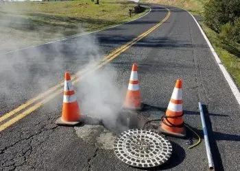 A road with steam rising from a manhole, traffic cones around it, and a pole lying nearby.