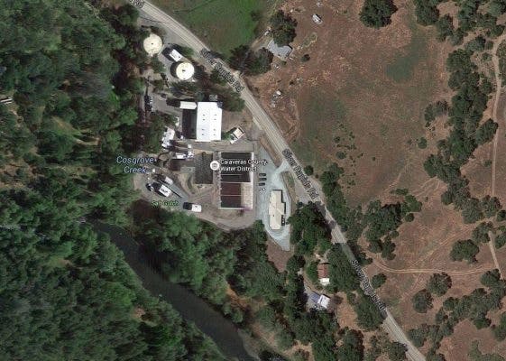 Aerial view of a facility with buildings and satellite dishes near a creek and treeline, with surrounding open land.