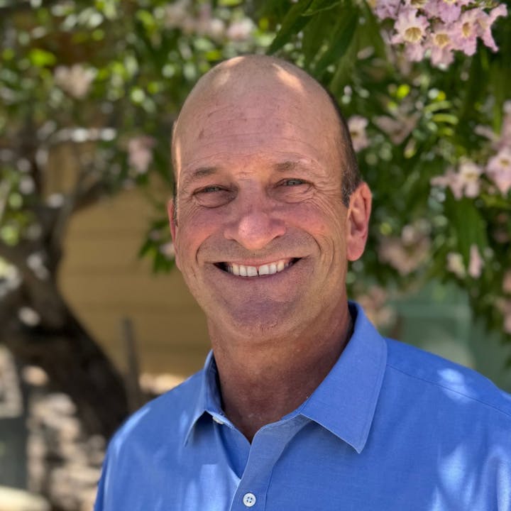 A smiling person in a blue shirt standing outdoors with a tree and pink flowers in the background.
