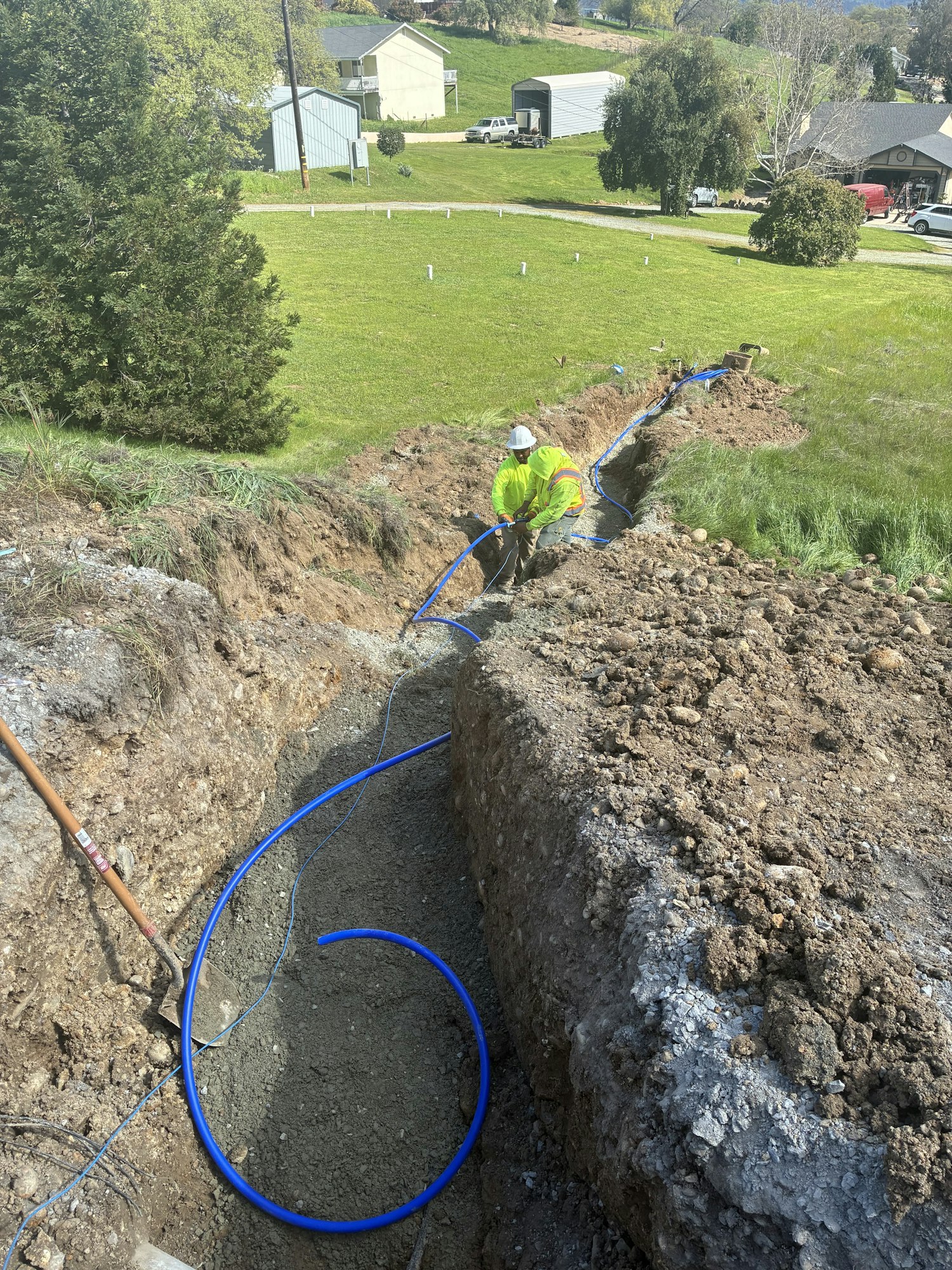 A worker in a yellow jacket is digging a trench and laying blue pipes in a grassy field near residential buildings.