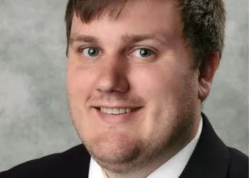 A headshot of a smiling man with short hair wearing a suit, with a blurred neutral background.