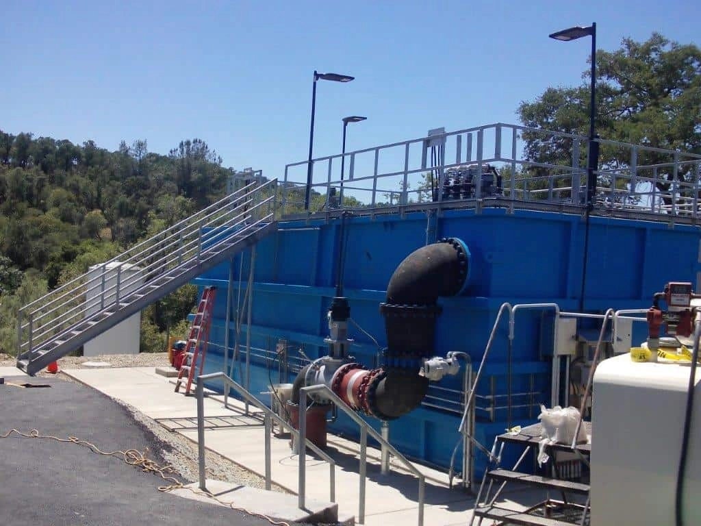 Blue industrial water treatment tanks, piping, and a staircase, with a forest background.