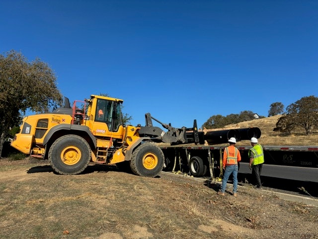 A large yellow loader is positioned near a truck, with two workers in safety vests directing the operation on a rural road.