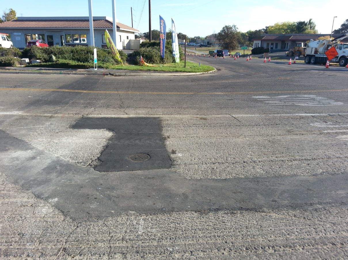 A road under repair with patched asphalt, traffic cones, utility vehicles, and a manhole cover, in a suburban setting.