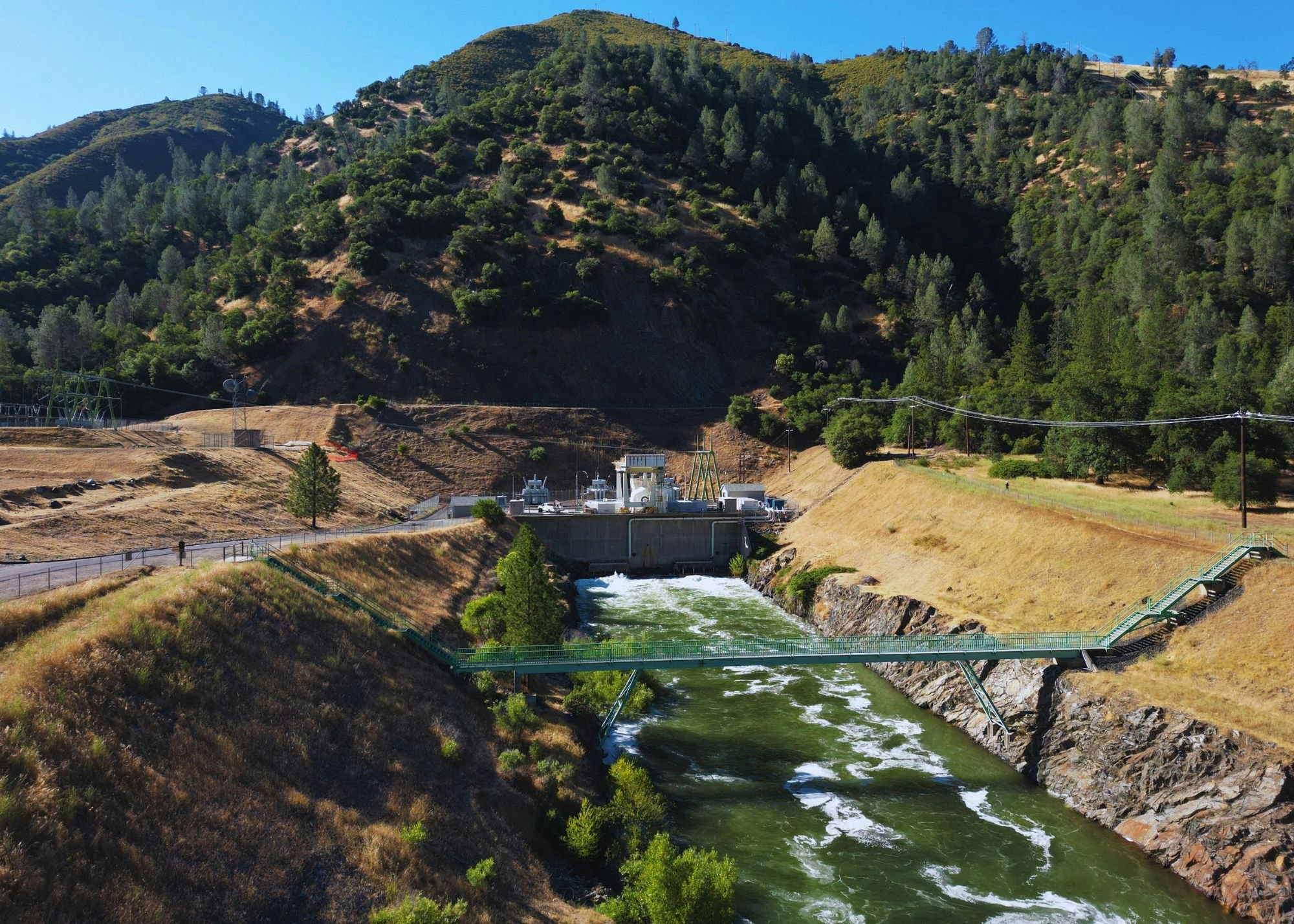 A dam with flowing water amidst green hills and trees, featuring a bridge and hydroelectric facility.