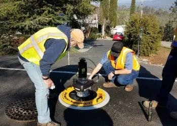 Two people in safety vests working with equipment on a street, possibly for utilities or maintenance.