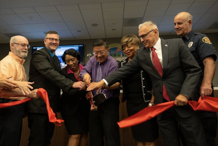 Group of people cutting a red ribbon, suggesting a ceremonial event or opening.