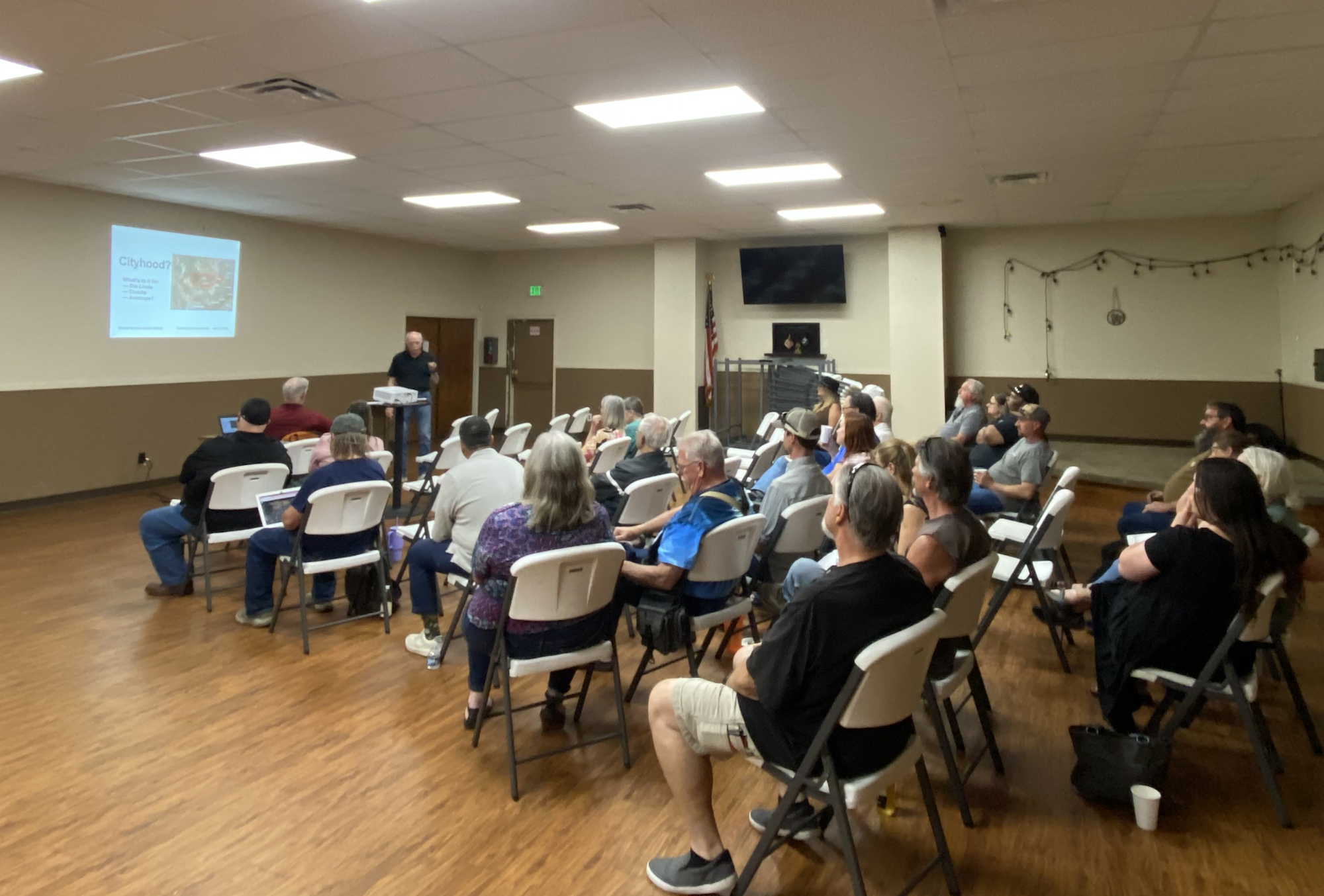 A speaker presenting to a group of seated people in a meeting room with a projected slide labeled "Cityhood?"