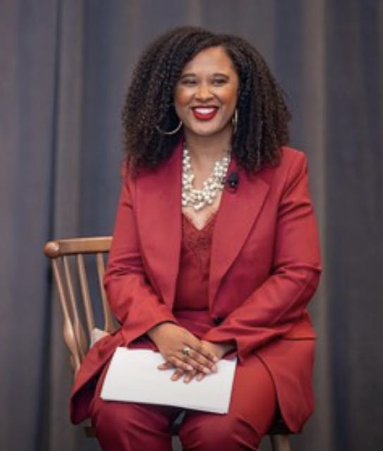 A woman in a red suit and pearl necklace, seated and smiling.