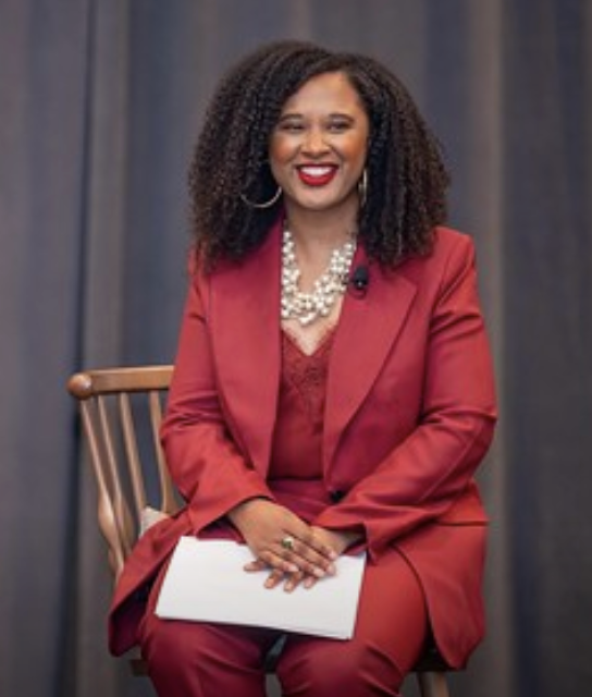 A woman in a red suit and pearl necklace, seated and smiling.