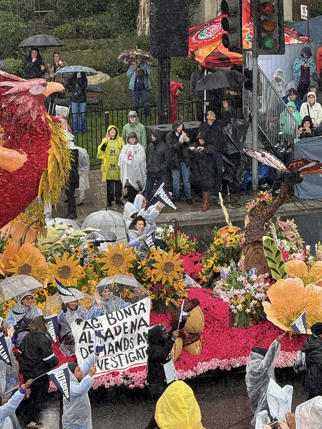 The image shows a colorful parade float with flowers, people holding umbrellas, and a protest sign in the rain.