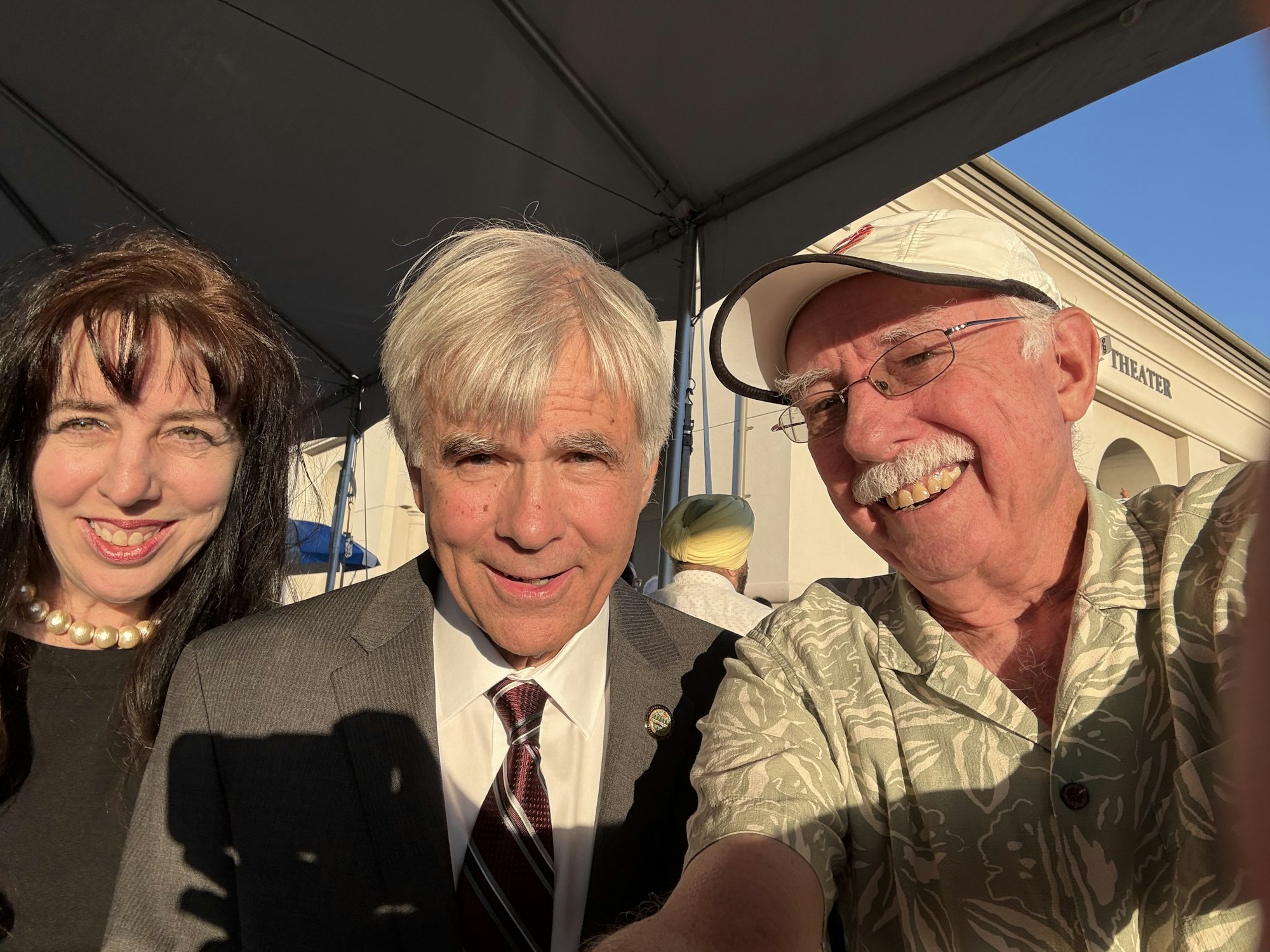 Three people smiling in sunlight, wearing formal and casual attire, with a canopy overhead.