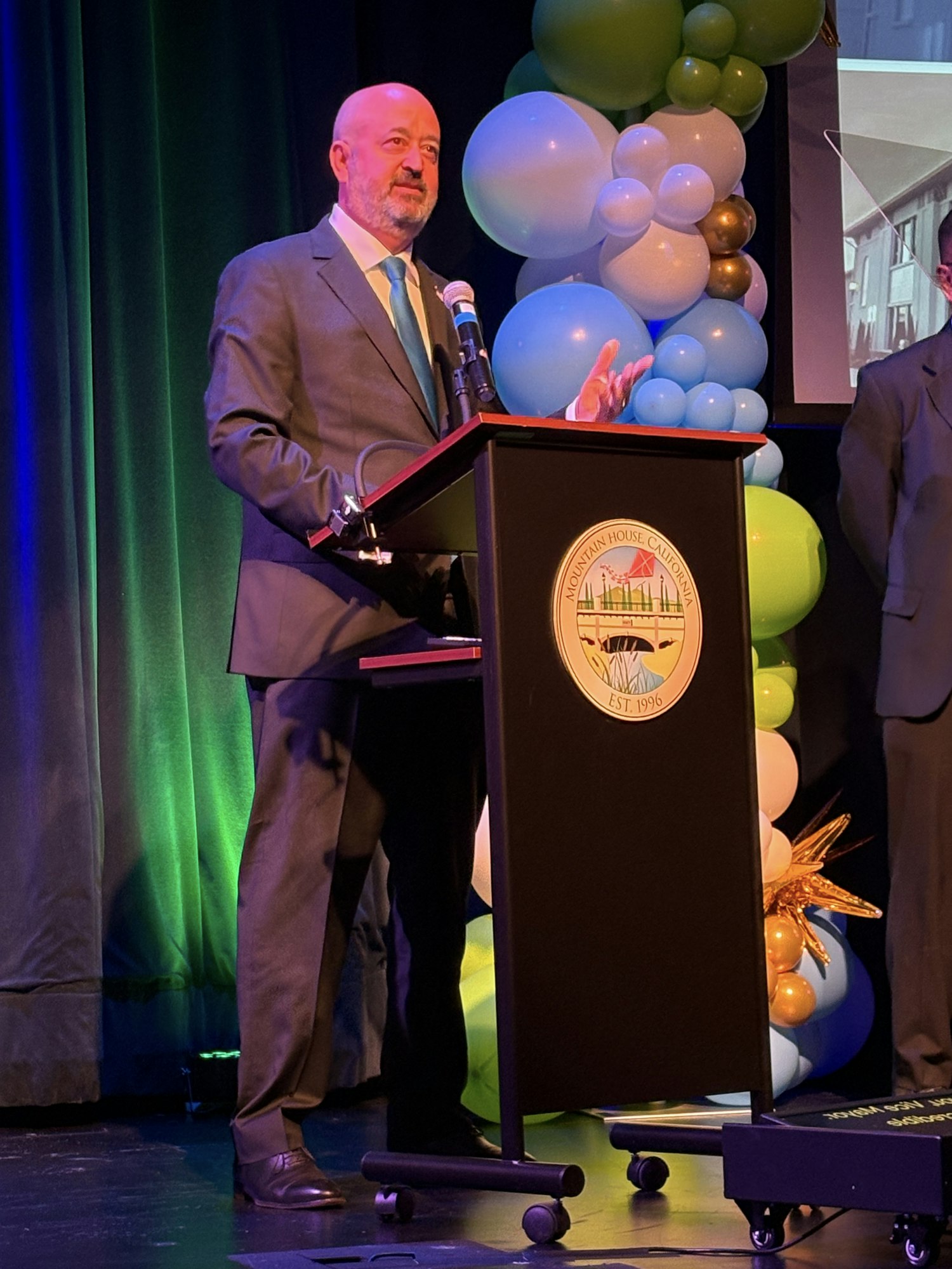 A person in a suit speaking at a podium with balloons in the background.