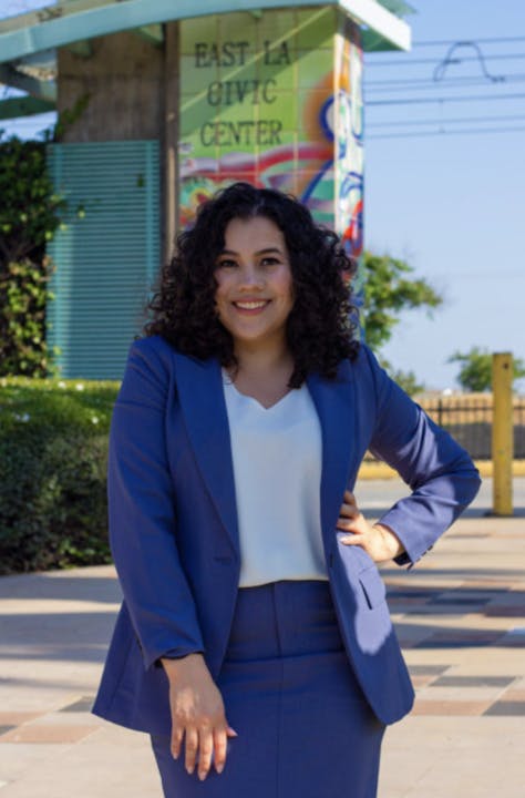 A smiling person in a blue suit standing in front of the East LA Civic Center with artistic elements in the background.