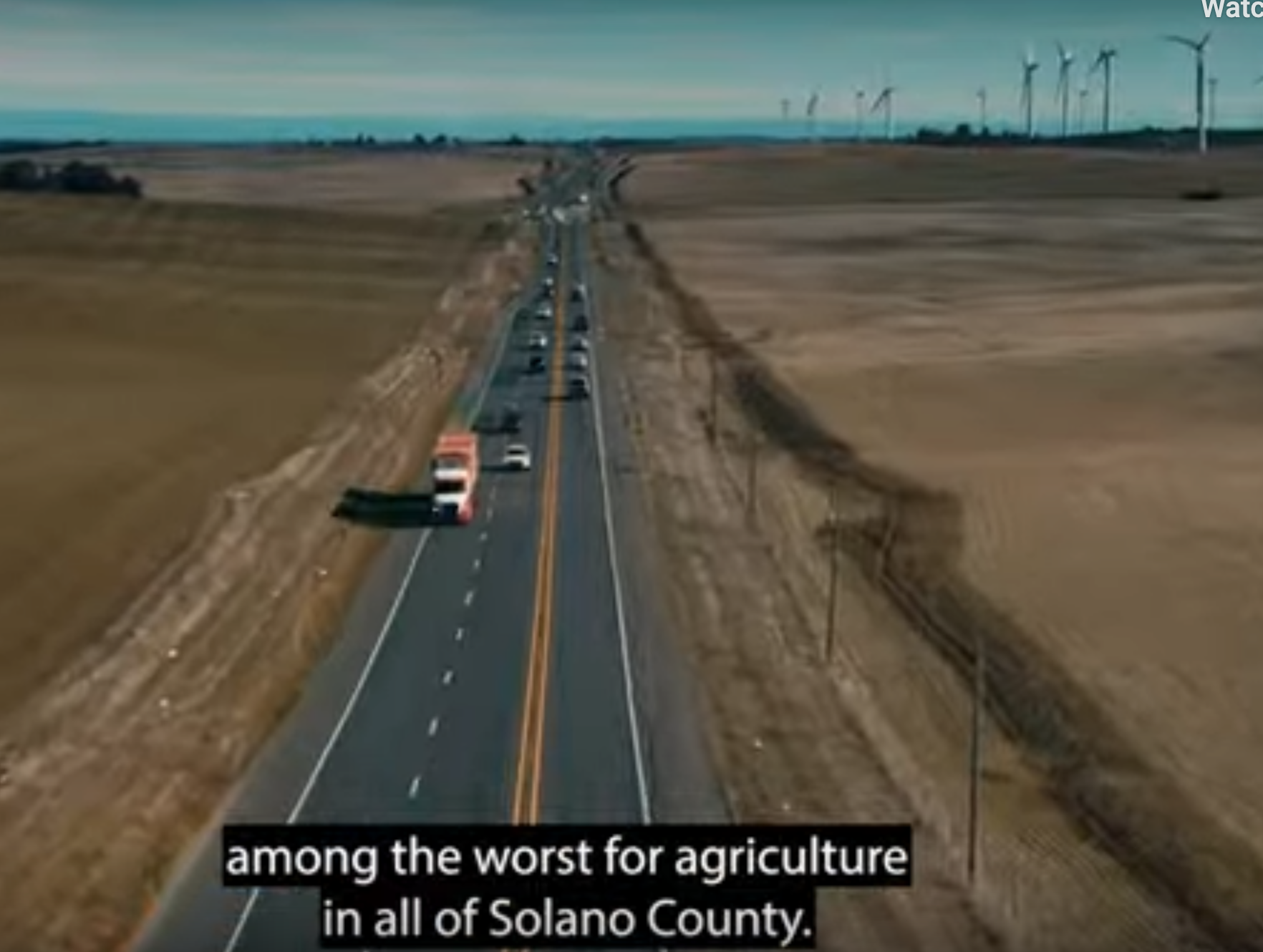 A highway alongside a barren field with wind turbines in the distance and a subtitle about agriculture in Solano County.